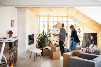 Bright living room with exposed brick, staircase, and a moving crew carrying boxes and furniture into the space, with cardboard boxes stacked near the sofa.