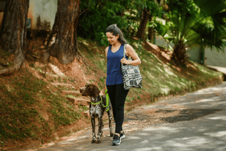 A person jogging on a winding road in a green, wooded area, wearing a blue patterned tank top, dark leggings, and a small crossbody bag.