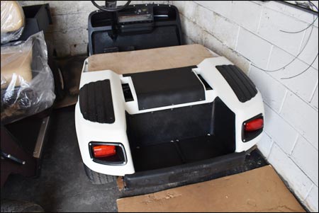 A white golf cart without a roof or seats is parked indoors next to a white brick wall, with cardboard pieces on the floor nearby.
