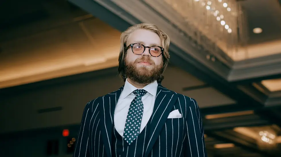 Man in a pinstripe suit with glasses and a beard standing in a formal indoor setting with chandeliers in the background