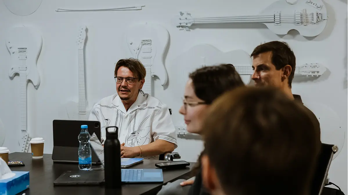 People in a meeting room with white guitar wall decorations, discussing around a table with laptops