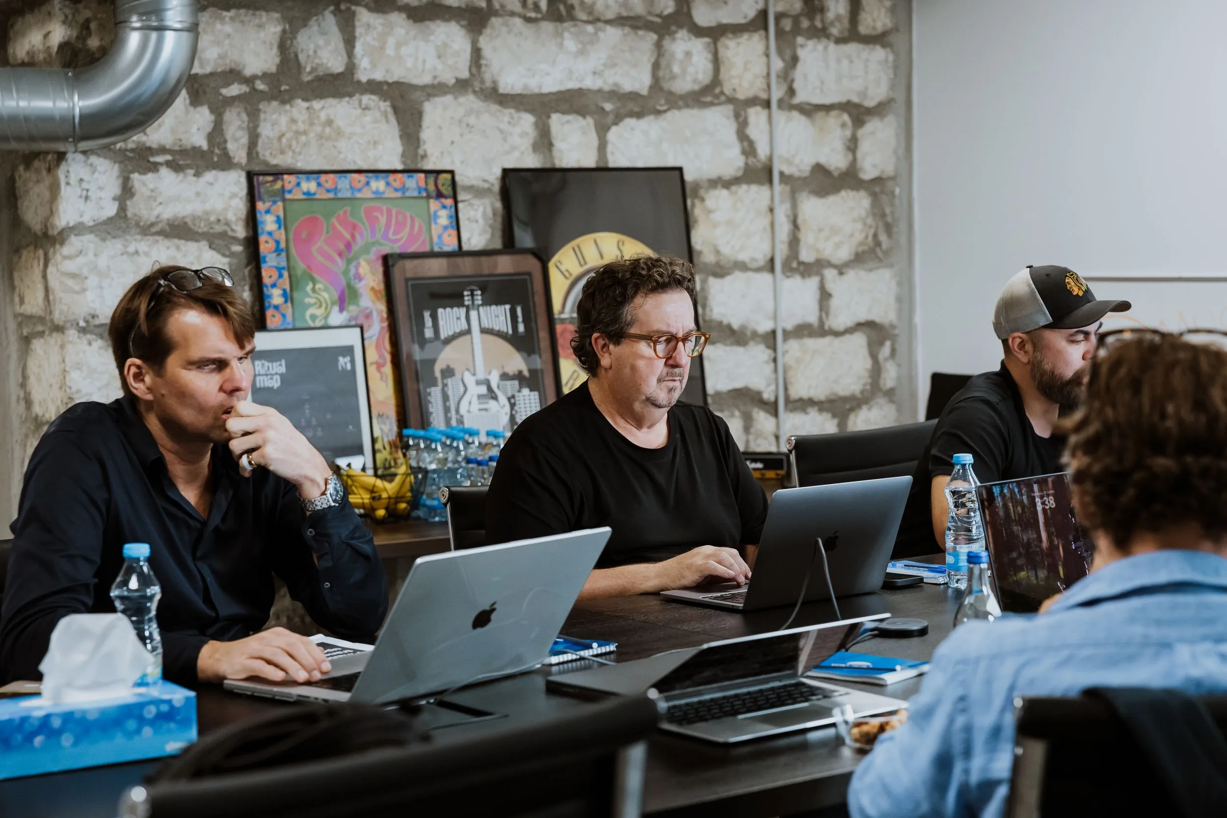 Group of people working together at a table with laptops in a modern office with stone walls and music posters