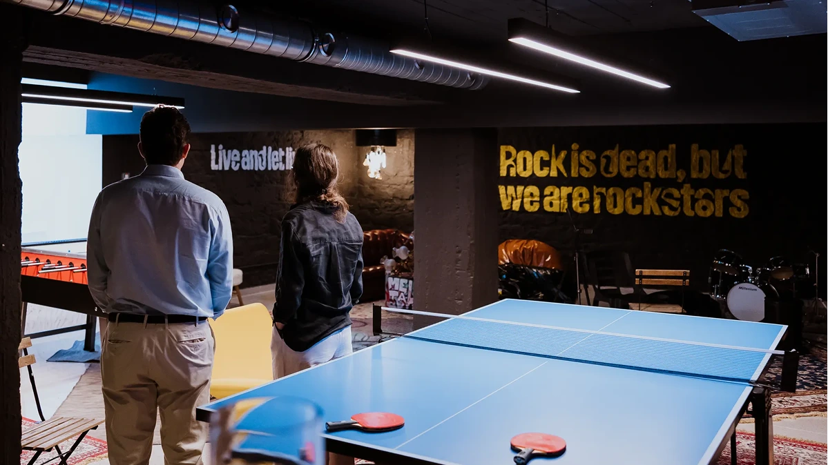 Two people standing in a recreation room with a ping-pong table and music-themed wall art.