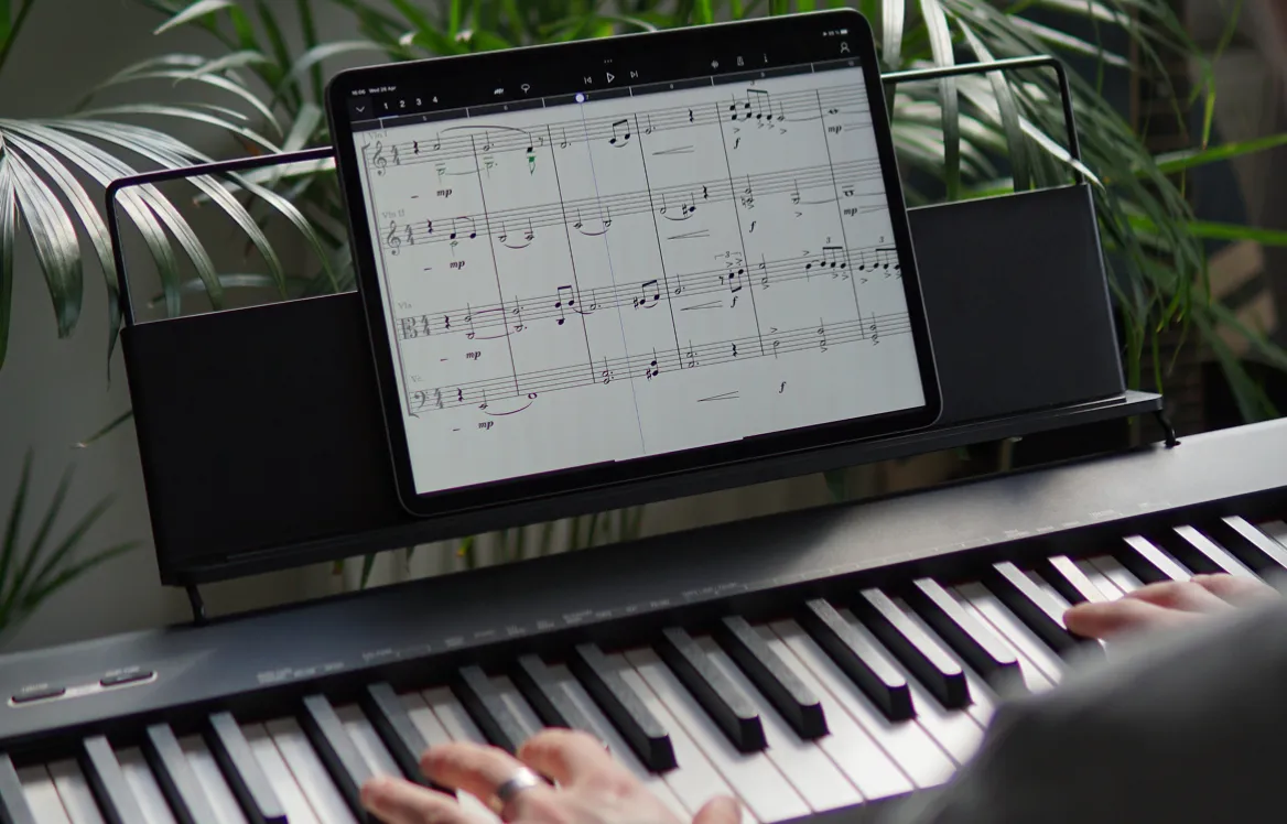 Hands playing a digital piano with a tablet displaying sheet music on the music stand, surrounded by green plants.