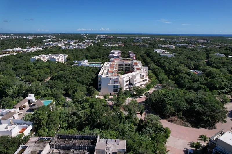 An aerial view of the Costa Caribe development in Tulum, Mexico, featuring a large, modern white building complex with multiple levels and balconies, surrounded by dense green jungle foliage under a clear blue sky, with other residential structure...