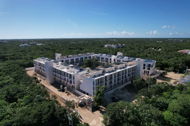 An aerial wide shot of the Natal condominium development in Tulum, Mexico, showcasing a large, square-shaped white building complex nestled within dense green jungle. The building features multiple levels, numerous windows, and solar panels visibl...