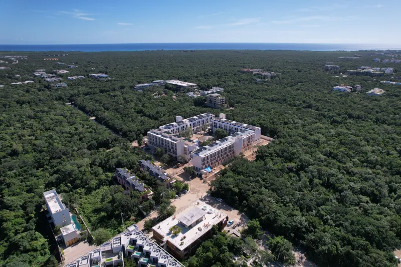 An aerial wide shot of the Natal condominium development in Tulum, Mexico, showcasing a large, square-shaped white building complex nestled within dense green jungle. The building features multiple levels, numerous windows, and solar panels visibl...