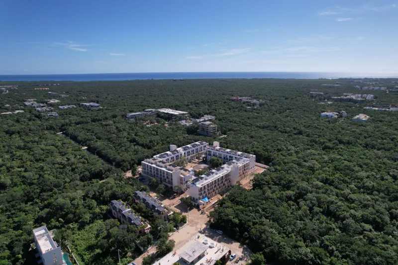 An aerial wide shot of the Natal condominium development in Tulum, Mexico, showcasing a large, square-shaped white building complex nestled within dense green jungle. The building features multiple levels, numerous windows, and solar panels visibl...
