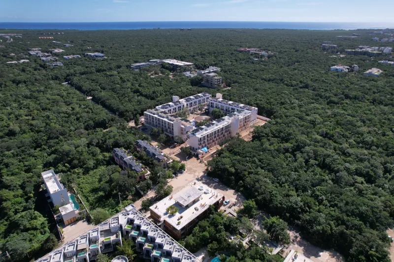 An aerial wide shot of the Natal condominium development in Tulum, Mexico, showcasing a large, square-shaped white building complex nestled within dense green jungle. The building features multiple levels, numerous windows, and solar panels visibl...
