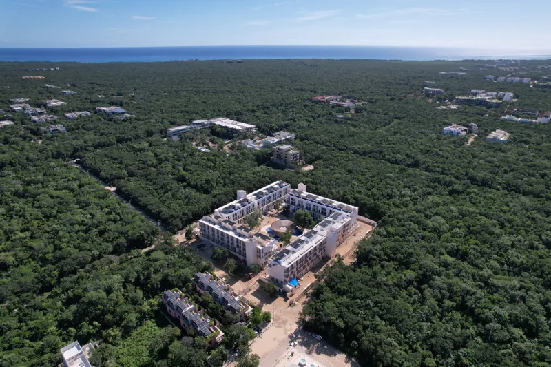 An aerial wide shot of the Natal condominium development in Tulum, Mexico, showcasing a large, square-shaped white building complex nestled within dense green jungle. The building features multiple levels, numerous windows, and solar panels visibl...
