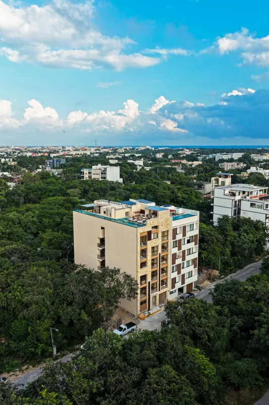 An aerial view of the Suutuk development in Playa del Carmen, showing a multi-story building with a light-colored facade, wooden accents, and a visible rooftop pool and lounge area. The building is surrounded by lush green trees, with other urban ...