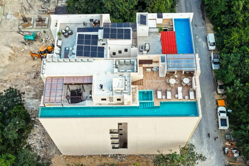 An aerial view of the Suutuk development in Playa del Carmen, showing a multi-story building with a light-colored facade, wooden accents, and a visible rooftop pool and lounge area. The building is surrounded by lush green trees, with other urban ...