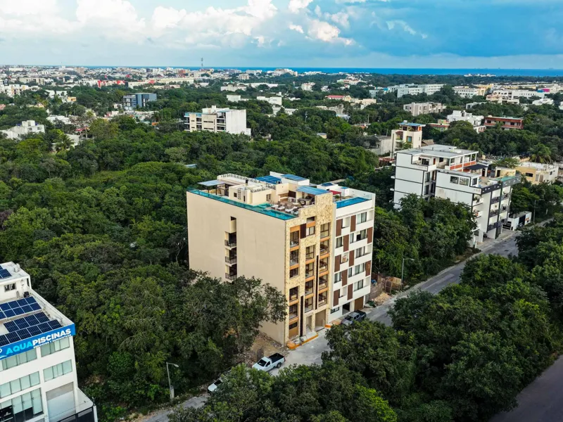 An aerial view of the Suutuk development in Playa del Carmen, showing a multi-story building with a light-colored facade, wooden accents, and a visible rooftop pool and lounge area. The building is surrounded by lush green trees, with other urban ...