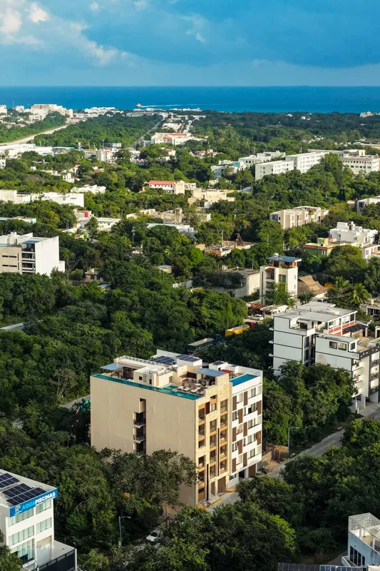 An aerial view of the Suutuk development in Playa del Carmen, showing a multi-story building with a light-colored facade, wooden accents, and a visible rooftop pool and lounge area. The building is surrounded by lush green trees, with other urban ...
