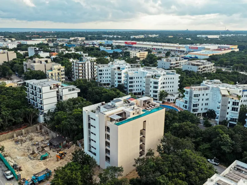 An aerial view of the Suutuk development in Playa del Carmen, showing a multi-story building with a light-colored facade, wooden accents, and a visible rooftop pool and lounge area. The building is surrounded by lush green trees, with other urban ...