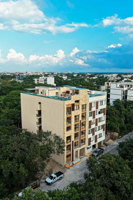 An aerial view of the Suutuk development in Playa del Carmen, showing a multi-story building with a light-colored facade, wooden accents, and a visible rooftop pool and lounge area. The building is surrounded by lush green trees, with other urban ...