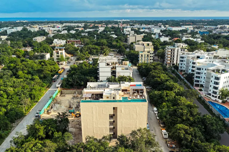 An aerial view of the Suutuk development in Playa del Carmen, showing a multi-story building with a light-colored facade, wooden accents, and a visible rooftop pool and lounge area. The building is surrounded by lush green trees, with other urban ...