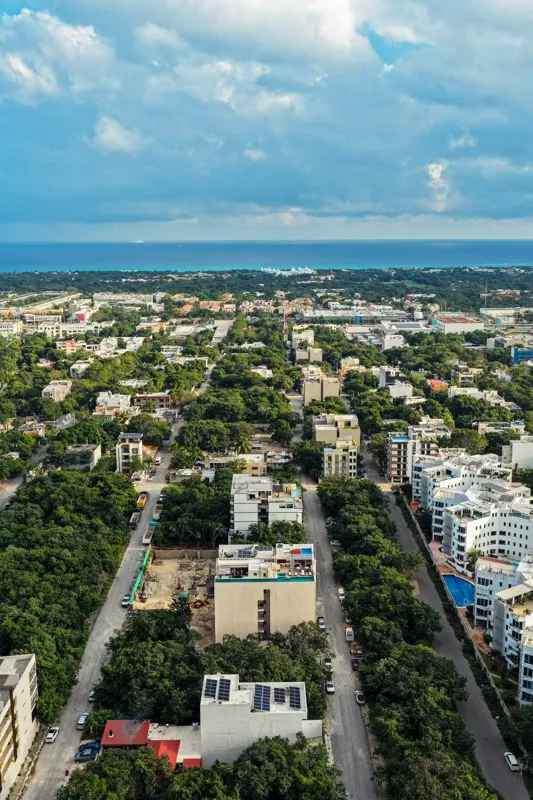 An aerial view of the Suutuk development in Playa del Carmen, showing a multi-story building with a light-colored facade, wooden accents, and a visible rooftop pool and lounge area. The building is surrounded by lush green trees, with other urban ...