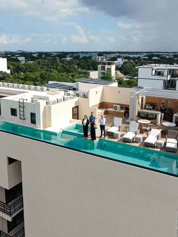 Aerial view of Suutuk's completed rooftop amenities, featuring a long turquoise infinity pool, a smaller jacuzzi, modern lounge furniture, a bar area, and solar panels, surrounded by lush green trees and other buildings in Playa del Carmen.