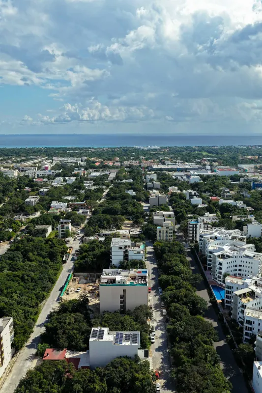 Aerial view of Suutuk's completed rooftop amenities, featuring a long turquoise infinity pool, a smaller jacuzzi, modern lounge furniture, a bar area, and solar panels, surrounded by lush green trees and other buildings in Playa del Carmen.