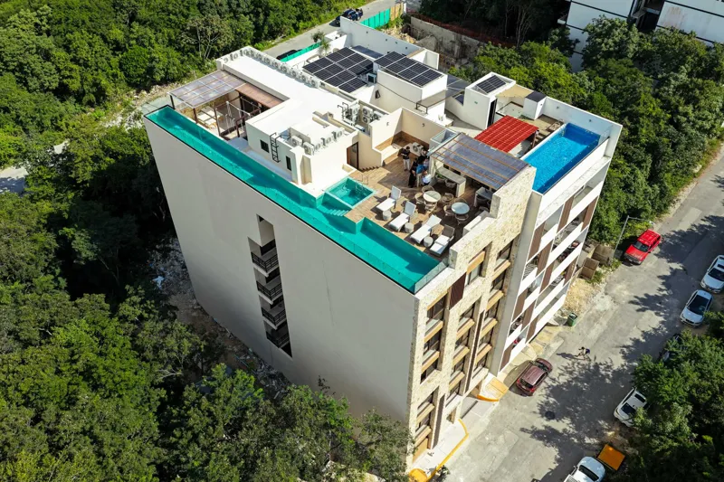 Aerial view of Suutuk's completed rooftop amenities, featuring a long turquoise infinity pool, a smaller jacuzzi, modern lounge furniture, a bar area, and solar panels, surrounded by lush green trees and other buildings in Playa del Carmen.