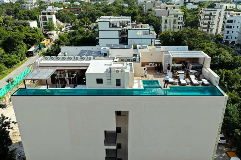 Aerial view of Suutuk's completed rooftop amenities, featuring a long turquoise infinity pool, a smaller jacuzzi, modern lounge furniture, a bar area, and solar panels, surrounded by lush green trees and other buildings in Playa del Carmen.