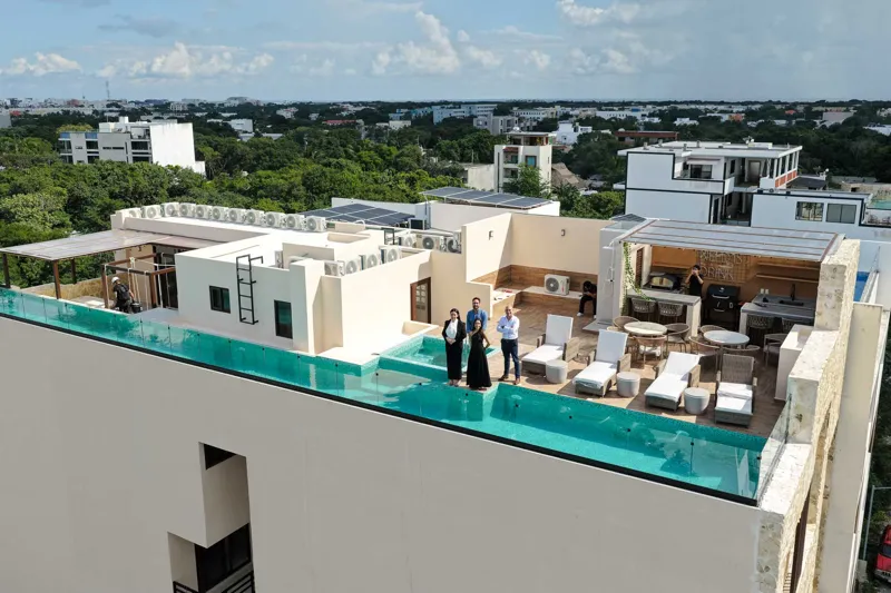 Aerial view of Suutuk's completed rooftop amenities, featuring a long turquoise infinity pool, a smaller jacuzzi, modern lounge furniture, a bar area, and solar panels, surrounded by lush green trees and other buildings in Playa del Carmen.