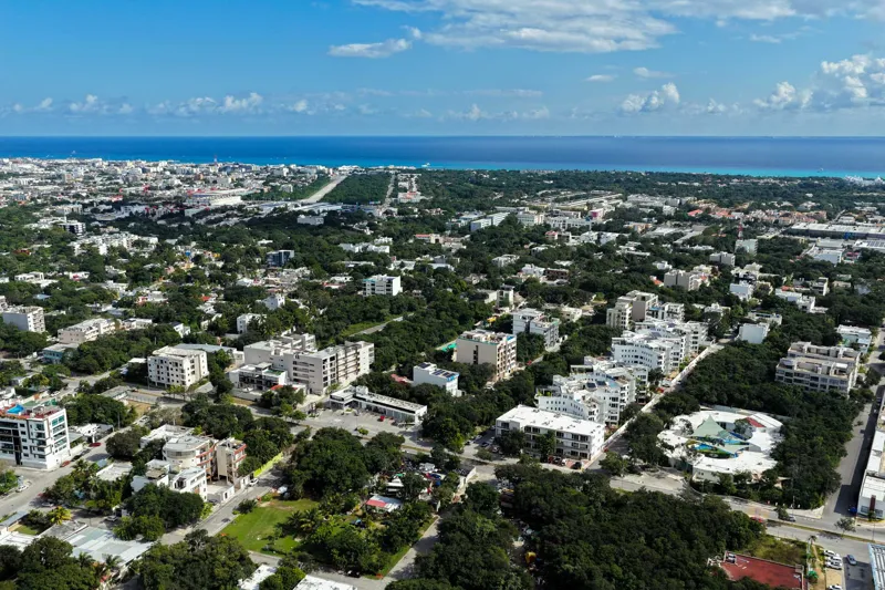 Aerial view of Suutuk's completed rooftop amenities, featuring a long turquoise infinity pool, a smaller jacuzzi, modern lounge furniture, a bar area, and solar panels, surrounded by lush green trees and other buildings in Playa del Carmen.