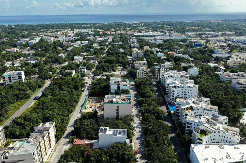 Aerial view of Suutuk's completed rooftop amenities, featuring a long turquoise infinity pool, a smaller jacuzzi, modern lounge furniture, a bar area, and solar panels, surrounded by lush green trees and other buildings in Playa del Carmen.