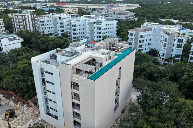 Aerial view of Suutuk's completed rooftop amenities, featuring a long turquoise infinity pool, a smaller jacuzzi, modern lounge furniture, a bar area, and solar panels, surrounded by lush green trees and other buildings in Playa del Carmen.