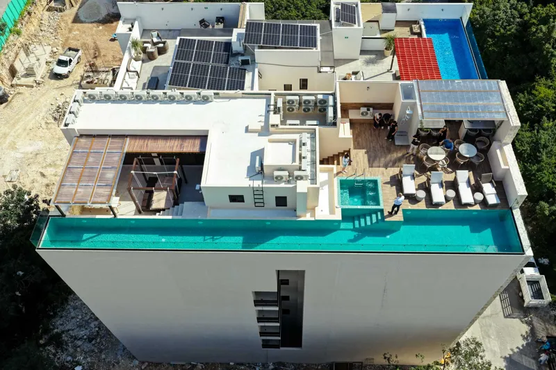 Aerial view of Suutuk's completed rooftop amenities, featuring a long turquoise infinity pool, a smaller jacuzzi, modern lounge furniture, a bar area, and solar panels, surrounded by lush green trees and other buildings in Playa del Carmen.