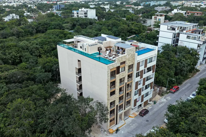 Aerial view of Suutuk's completed rooftop amenities, featuring a long turquoise infinity pool, a smaller jacuzzi, modern lounge furniture, a bar area, and solar panels, surrounded by lush green trees and other buildings in Playa del Carmen.