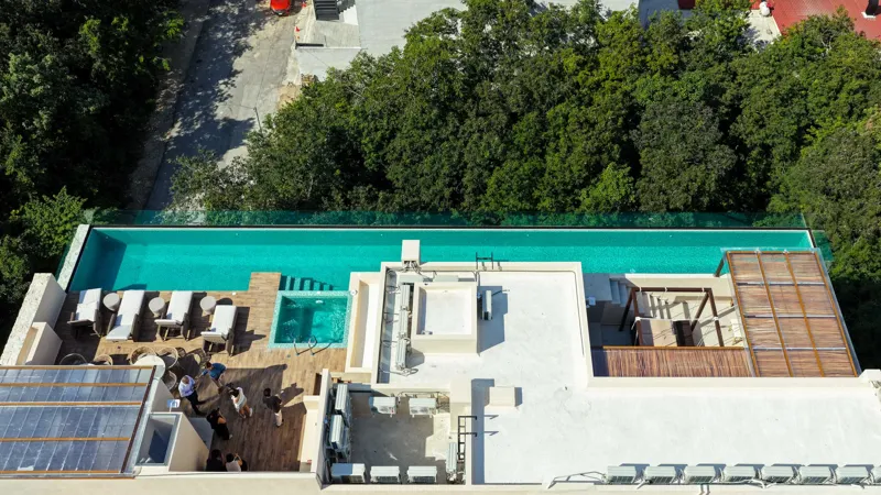 Aerial view of Suutuk's completed rooftop amenities, featuring a long turquoise infinity pool, a smaller jacuzzi, modern lounge furniture, a bar area, and solar panels, surrounded by lush green trees and other buildings in Playa del Carmen.
