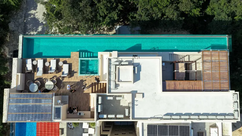Aerial view of Suutuk's completed rooftop amenities, featuring a long turquoise infinity pool, a smaller jacuzzi, modern lounge furniture, a bar area, and solar panels, surrounded by lush green trees and other buildings in Playa del Carmen.