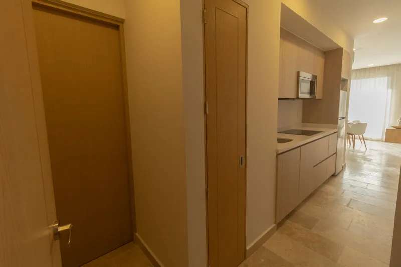 A fully furnished bedroom in a Vibbe model unit, featuring a large bed with a light wood frame, a wall-mounted television, a black ceiling fan, and light-colored walls adorned with decorative woven plates. A built-in light wood closet is visible o...