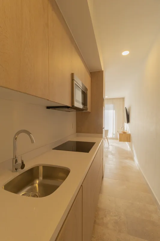 A fully furnished bedroom in a Vibbe model unit, featuring a large bed with a light wood frame, a wall-mounted television, a black ceiling fan, and light-colored walls adorned with decorative woven plates. A built-in light wood closet is visible o...