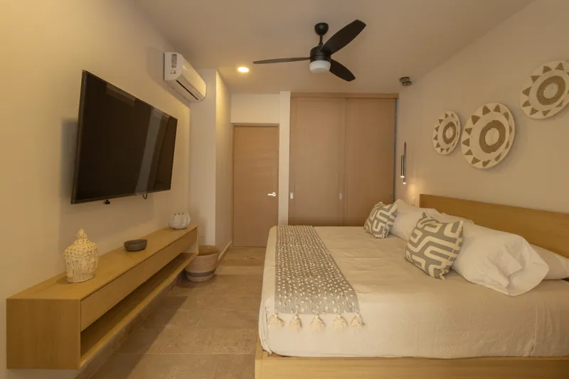 A fully furnished bedroom in a Vibbe model unit, featuring a large bed with a light wood frame, a wall-mounted television, a black ceiling fan, and light-colored walls adorned with decorative woven plates. A built-in light wood closet is visible o...