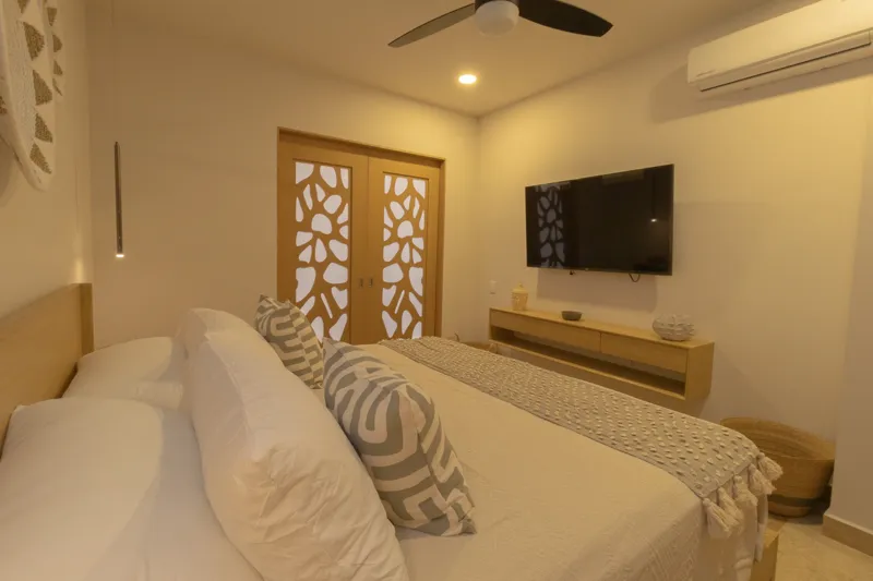 A fully furnished bedroom in a Vibbe model unit, featuring a large bed with a light wood frame, a wall-mounted television, a black ceiling fan, and light-colored walls adorned with decorative woven plates. A built-in light wood closet is visible o...