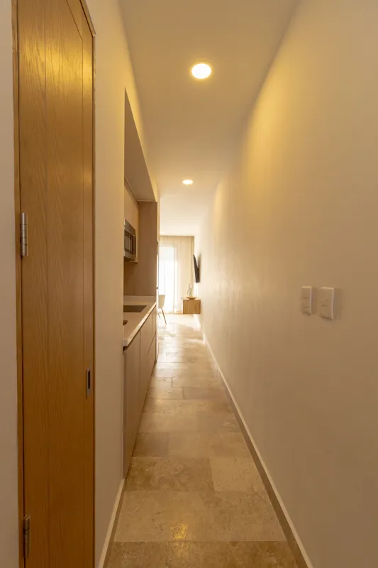 A fully furnished bedroom in a Vibbe model unit, featuring a large bed with a light wood frame, a wall-mounted television, a black ceiling fan, and light-colored walls adorned with decorative woven plates. A built-in light wood closet is visible o...