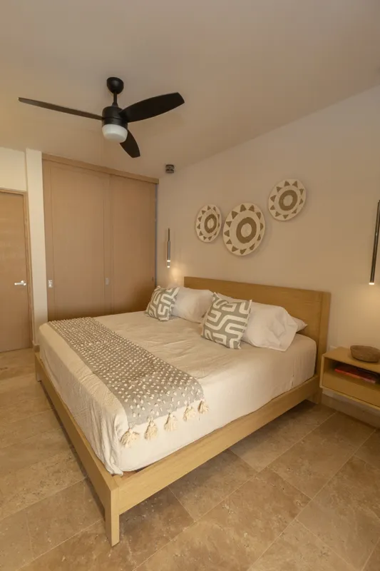 A fully furnished bedroom in a Vibbe model unit, featuring a large bed with a light wood frame, a wall-mounted television, a black ceiling fan, and light-colored walls adorned with decorative woven plates. A built-in light wood closet is visible o...
