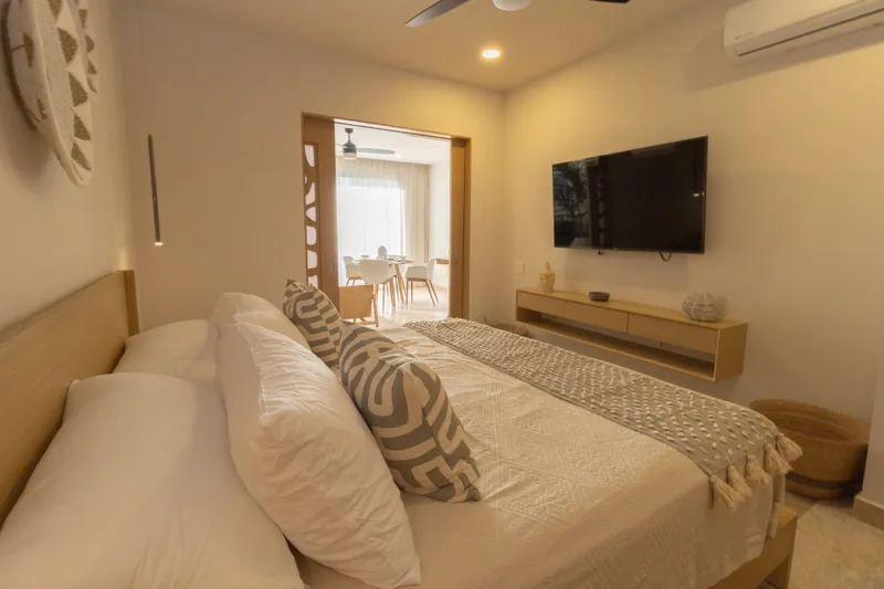 A fully furnished bedroom in a Vibbe model unit, featuring a large bed with a light wood frame, a wall-mounted television, a black ceiling fan, and light-colored walls adorned with decorative woven plates. A built-in light wood closet is visible o...