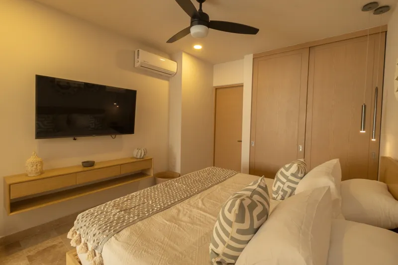 A fully furnished bedroom in a Vibbe model unit, featuring a large bed with a light wood frame, a wall-mounted television, a black ceiling fan, and light-colored walls adorned with decorative woven plates. A built-in light wood closet is visible o...