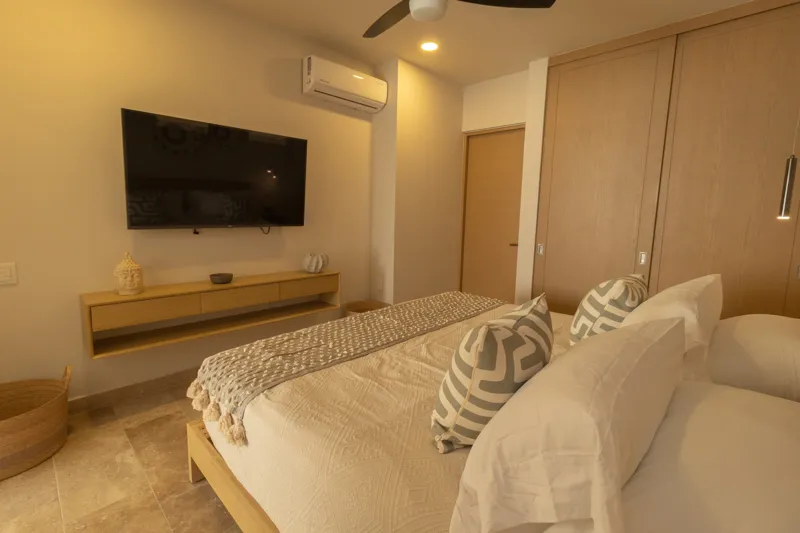 A fully furnished bedroom in a Vibbe model unit, featuring a large bed with a light wood frame, a wall-mounted television, a black ceiling fan, and light-colored walls adorned with decorative woven plates. A built-in light wood closet is visible o...