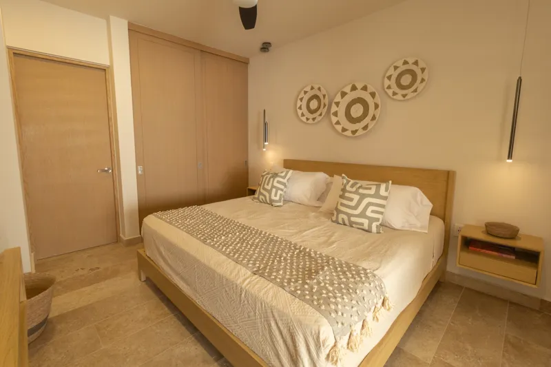 A fully furnished bedroom in a Vibbe model unit, featuring a large bed with a light wood frame, a wall-mounted television, a black ceiling fan, and light-colored walls adorned with decorative woven plates. A built-in light wood closet is visible o...