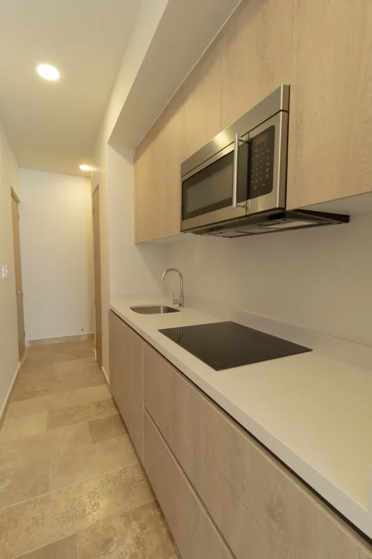 A fully furnished bedroom in a Vibbe model unit, featuring a large bed with a light wood frame, a wall-mounted television, a black ceiling fan, and light-colored walls adorned with decorative woven plates. A built-in light wood closet is visible o...