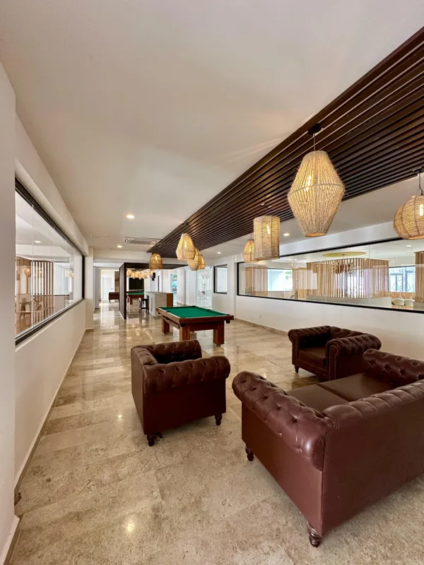 A vibrant rooftop pool bar at TownCenter Zama, featuring a sparkling blue swimming pool, a bar counter with four wooden stools, and modern pendant lights, all set against a clear blue sky with lush green foliage in the background, showcasing a rea...