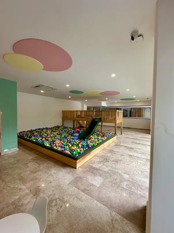 A vibrant rooftop pool bar at TownCenter Zama, featuring a sparkling blue swimming pool, a bar counter with four wooden stools, and modern pendant lights, all set against a clear blue sky with lush green foliage in the background, showcasing a rea...