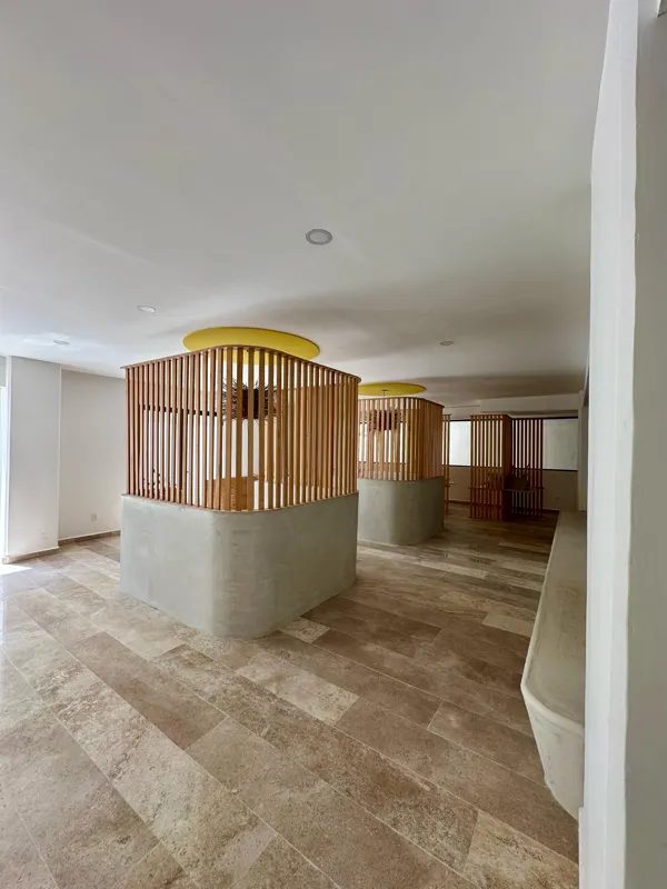 A vibrant rooftop pool bar at TownCenter Zama, featuring a sparkling blue swimming pool, a bar counter with four wooden stools, and modern pendant lights, all set against a clear blue sky with lush green foliage in the background, showcasing a rea...