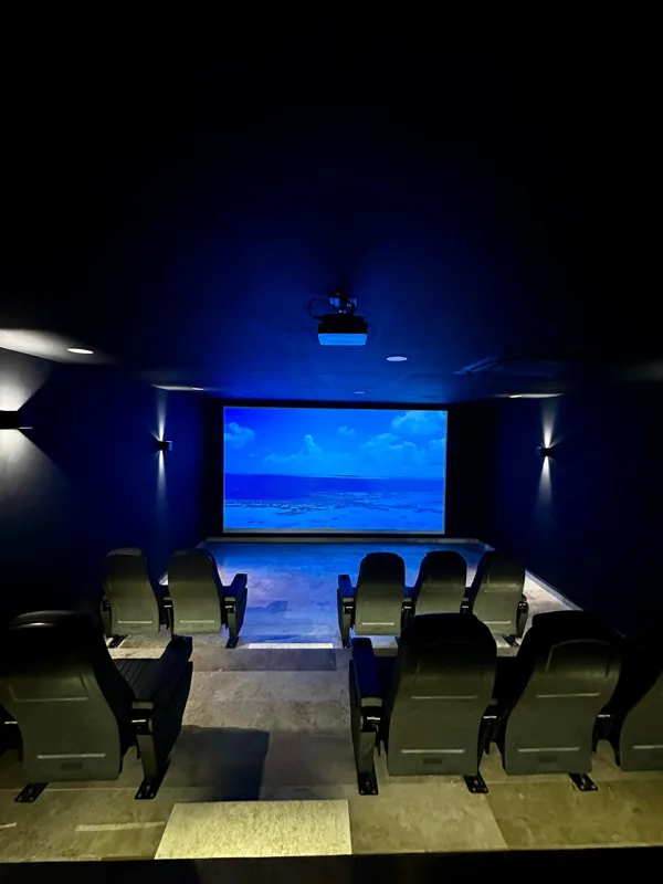 A vibrant rooftop pool bar at TownCenter Zama, featuring a sparkling blue swimming pool, a bar counter with four wooden stools, and modern pendant lights, all set against a clear blue sky with lush green foliage in the background, showcasing a rea...