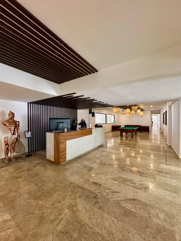 A vibrant rooftop pool bar at TownCenter Zama, featuring a sparkling blue swimming pool, a bar counter with four wooden stools, and modern pendant lights, all set against a clear blue sky with lush green foliage in the background, showcasing a rea...